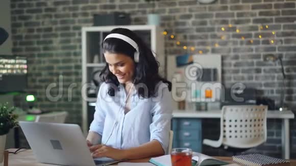 会玩的女孩跳舞在耳机里欣赏音乐在办公室里使用笔记本电脑视频的预览图