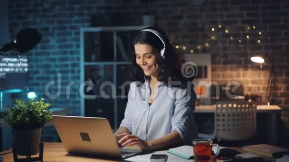 女人在笔记本电脑上打字晚上在黑暗的办公室里用耳机听音乐视频的预览图
