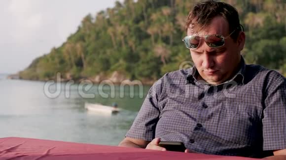 一位英俊的年轻商人戴着太阳镜在海滩咖啡馆里背景是海景使用手机视频的预览图