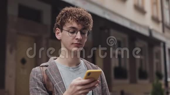 戴着夹克戴着眼镜戴着红色卷发的年轻嬉皮士用手机打字留言的肖像视频的预览图