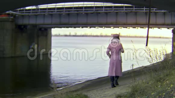 后景一个孤独美丽的女人穿着温暖的秋装走在河边的桥下库存数据视频的预览图