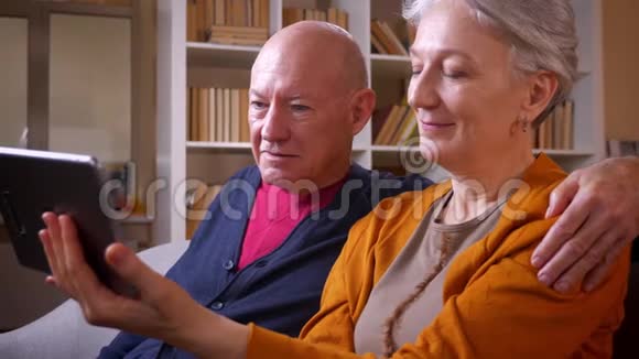 高级白种人配偶的侧写镜头看着平板电脑坐在客厅的沙发上被逗乐视频的预览图