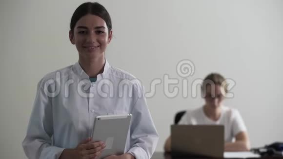 描绘喜悦的年轻职业女医生拿着笔记本电脑看着相机在背景中坐着一个视频的预览图