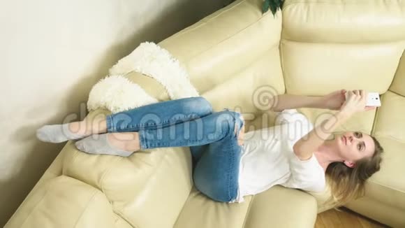 可爱的金发女孩躺在沙发上用手机自拍视频的预览图