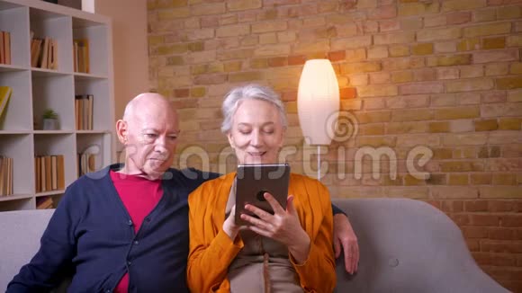 老白种人夫妇兴高采烈地看着平板电脑坐在沙发上在客厅里互相拥抱视频的预览图