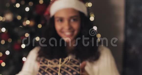 圣诞帽里漂亮的微笑女孩把圣诞礼物盒送给相机站在装饰灯光下视频的预览图