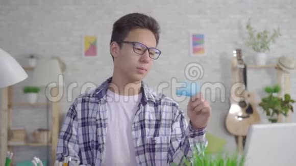 一个体贴的亚洲年轻人戴着时髦的眼镜在笔记本电脑上工作在房子的客厅里拿着一张银行卡视频的预览图