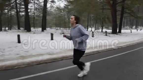 年轻迷人的白种人女孩在冬天带着耳机在雪地公园跑步前面跟着射击慢动作视频的预览图