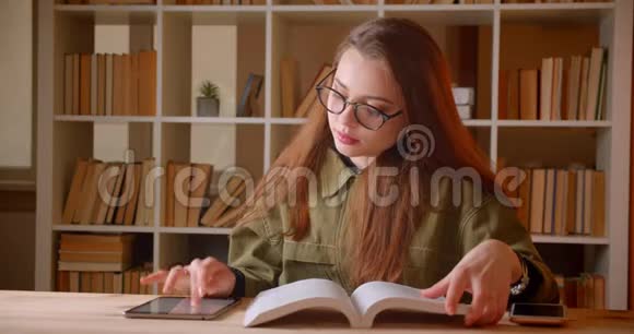 年轻女学生阅读书籍和平板电脑搜索信息的肖像在图书馆变成了相机视频的预览图