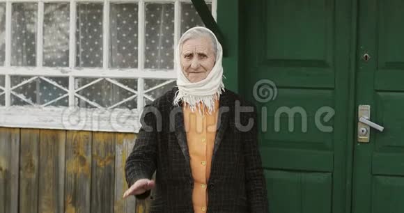 一位戴着围巾的皱纹很深的老妇人站着在一所乡村房子的背景上挥动她的手视频的预览图