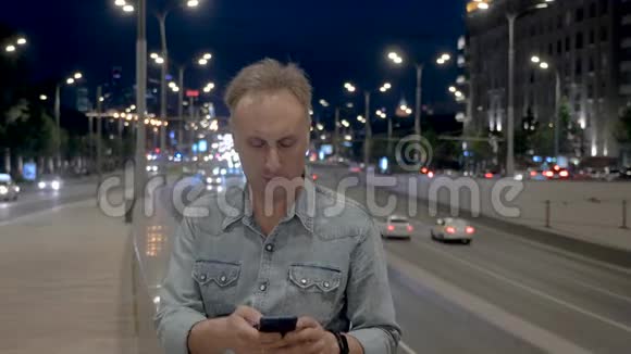 中年男子在晚间城市的女儿墙上使用智能手机视频的预览图