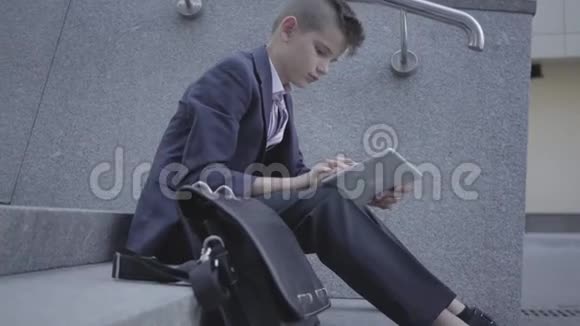 穿着西装的小男孩坐在街上的楼梯上在平板电脑上打字作为成年人的孩子视频的预览图