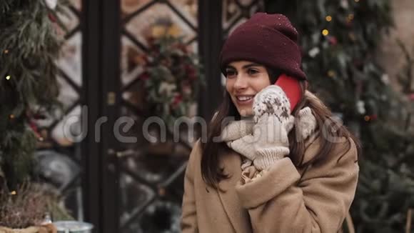 年轻的微笑迷人的女孩戴着冬帽和手套站在外面和一个朋友聊天打电话视频的预览图