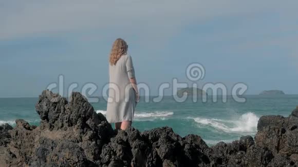 年轻女孩穿着灰色斗篷站在风暴波附近撞击岩石年轻女孩望着大海海浪拍打着视频的预览图