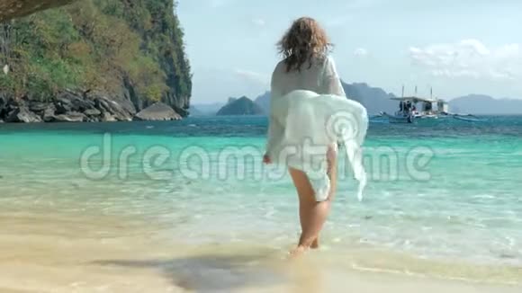 海滩上穿着蓝色斗篷的漂亮女孩的后景年轻迷人的金发女郎走在热带海滩上视频的预览图