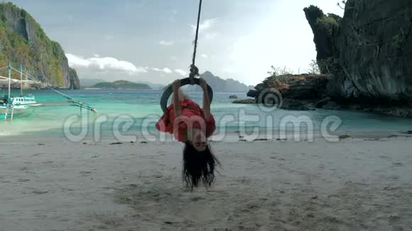 在阳光明媚的日子里穿着红色连衣裙的年轻女子在安静的沙滩上荡着绳子旅行概念巴拉望岛视频的预览图