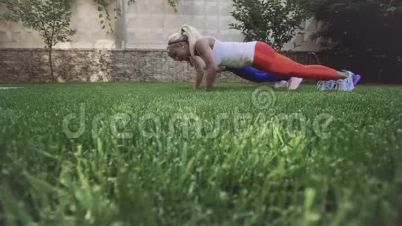 穿白色t恤的金发女孩在新鲜空气中的绿草上做运动锻炼视频的预览图
