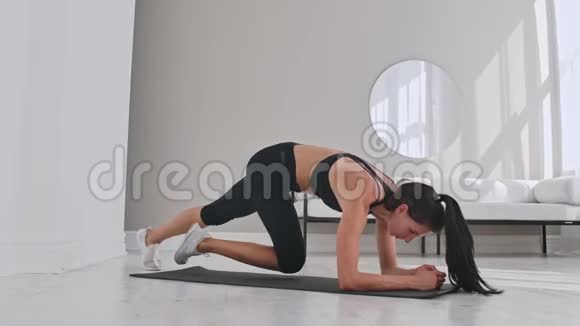 女性黑发健身教练在家里的垫子上展示木板膝盖到胸部的技术视频的预览图