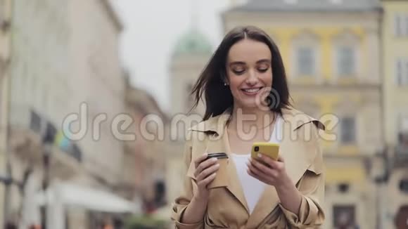 年轻漂亮的女孩喝着咖啡用她的智能手机微笑在城市街上打字视频的预览图