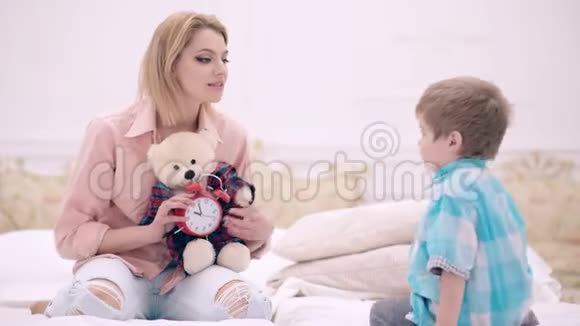年轻的妈妈带着可爱的小儿子和泰迪熊坐在家里的床上友好的家庭一位年轻的母亲坐在旁边视频的预览图