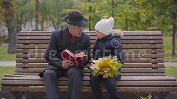 一位穿着经典服装的白种人成年男子和孙女坐在长凳上看书的画像视频的预览图