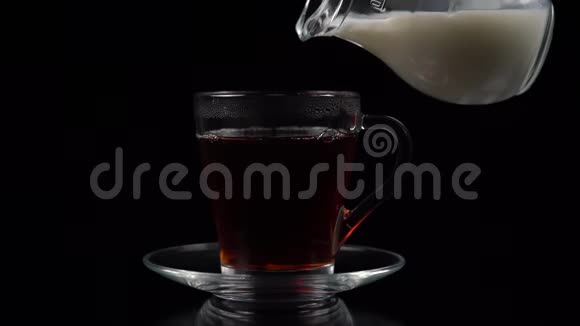 在黑茶中在玻璃茶杯和茶碟中倒入牛奶从玻璃壶中倒入黑色背景慢镜头拍摄视频的预览图