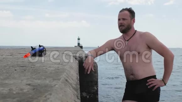 一位留着胡须的男游泳运动员站在一个混凝土码头上在灯塔的背景下在海里游泳4k4k视频视频的预览图