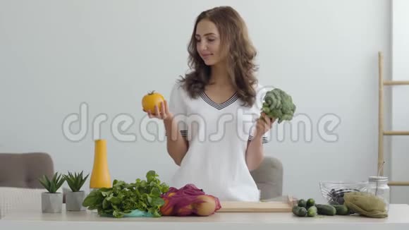 年轻微笑的女人拿着黄色的西红柿和西兰花站在现代厨房的桌子旁健康食品的概念视频的预览图