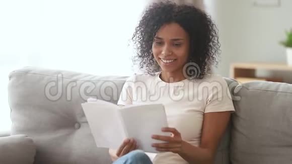 微笑着坐在沙发上看书的年轻非洲女人视频的预览图