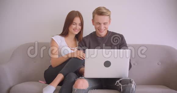 一对年轻可爱的白种人夫妇用笔记本电脑一起坐在公寓里的沙发上的特写照片视频的预览图