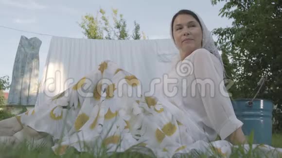 可爱的高级妇女头上戴着一条白色披肩坐在晾衣绳前面的草地上的花园里休息视频的预览图