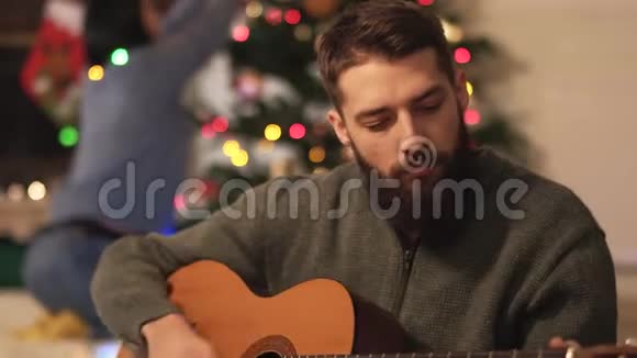 坐在圣诞树附近的地板上弹吉他唱歌的胡子男快乐的一对夫妇为圣诞节做准备视频的预览图