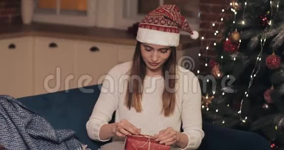 靠近迷人的女孩穿着毛衣和圣诞帽坐在圣诞树附近的沙发上最后敲定了一个大的视频的预览图