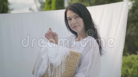 可爱的成熟女人留着长长的黑发手里拿着柳条篮子把白色的衣服挂在晾衣绳上视频的预览图