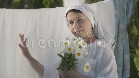 肖像高级妇女头上戴着白色披肩嗅着雏菊看着户外晾衣绳附近的摄像机视频的预览图