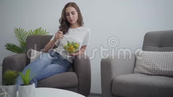 漂亮的年轻苗条的女人端着大碗和沙拉坐在扶手椅上享受健康的食物视频的预览图