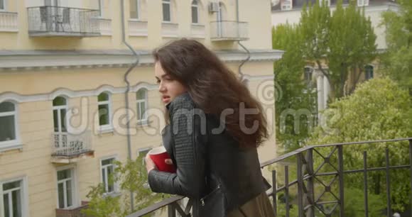 穿着皮夹克的漂亮黑发女孩从阳台上观看绿色城市景色喝着热茶是个梦幻般的女孩视频的预览图