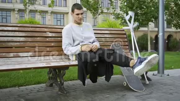 年轻人坐在公园的长凳上一边用手机听音乐然后拿起拐杖和滑板视频的预览图