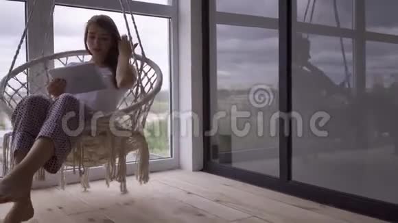 苗条可爱的年轻女子在一张挂着的椅子上摆动同时在大的落地到天花板的窗户上使用她的平板电脑自由职业者视频的预览图