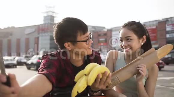 迷人的30多岁亚洲男人和女人微笑着拿着香蕉和三明治滚筒在大商店附近自拍视频的预览图