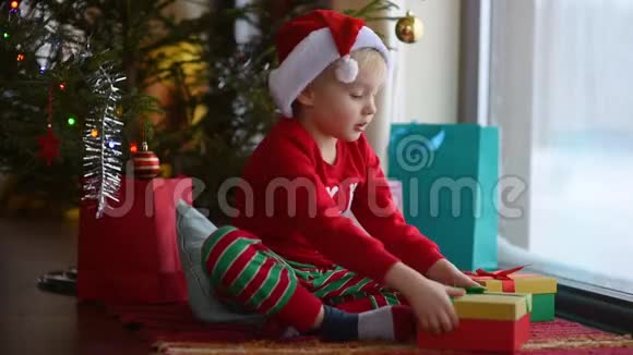 可爱的小男孩戴着圣诞老人的帽子打开圣诞礼物圣诞早晨快乐的孩子视频的预览图