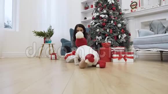 美丽的年轻妈妈坐在圣诞树附近的地板上给她那有趣的蹒跚学步的红球视频的预览图