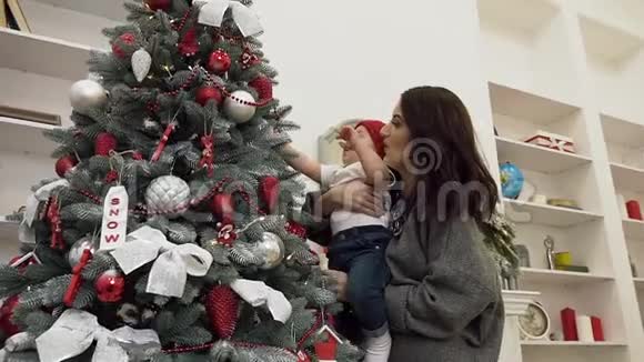 快乐的母亲让她不可思议的可爱的蹒跚学步的孩子触摸装饰精美的圣诞树视频的预览图