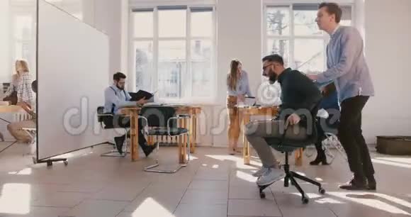 多利侧景拍摄疯狂有趣的商人骑着办公椅沿着繁忙的工作场所大厅庆祝成就视频的预览图