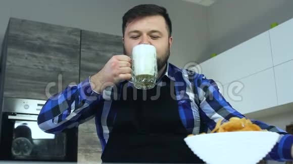 胖子坐在餐桌旁的厨房里喝着杯子里的泡沫啤酒一个穿着围裙的男人坐在桌子旁视频的预览图