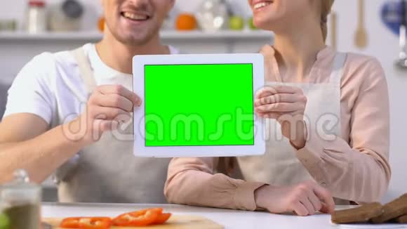 穿着围裙微笑的一对夫妇在相机上显示绿色屏幕平板电脑应用程序视频的预览图