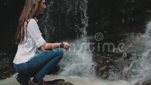 女孩徒步旅行者蹲在热带森林瀑布附近视频的预览图