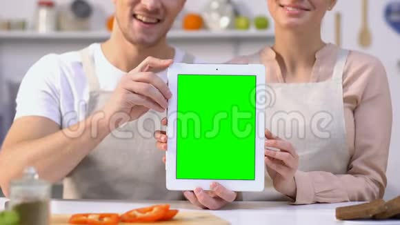 穿着围裙手里拿着绿色屏幕平板电脑坐在厨房收据上的一对快乐的夫妇视频的预览图