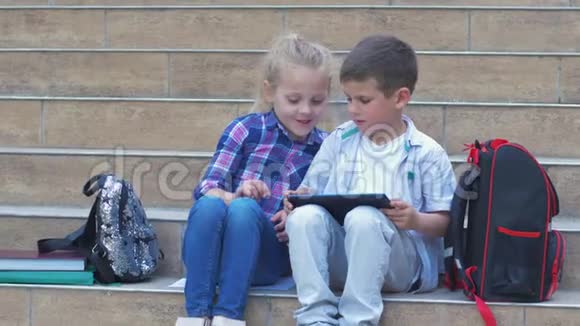 在户外课间休息时现代学龄儿童小女孩和小男孩在背包附近的台阶上使用数字平板电脑视频的预览图