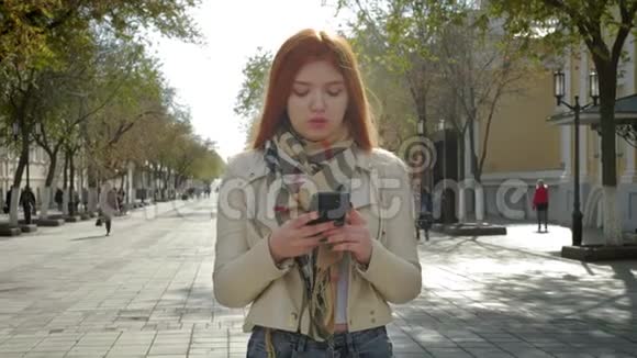 一个年轻的女孩站在一条城市街道上拿着智能手机输入短信与朋友聊天视频的预览图
